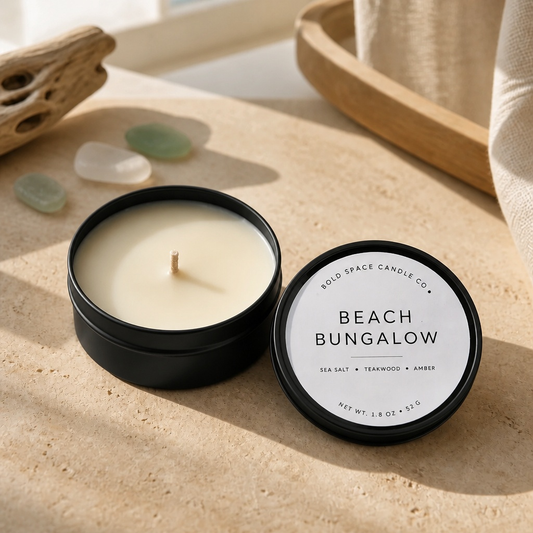 A black metal candle tin with its lid placed beside it on a beige stone surface. The candle wax is visible and the lid label reads 'BEACH BUNGALOW' with smaller text 'GOLD SPACE CANDLE CO.