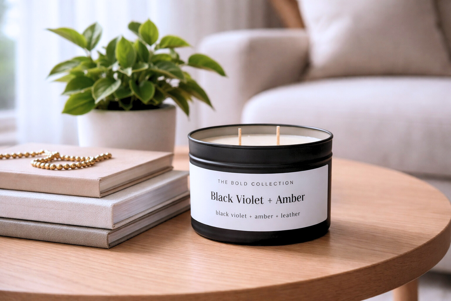 Candle labeled 'Black Violet + Amber' on a table with books and a plant in the background
