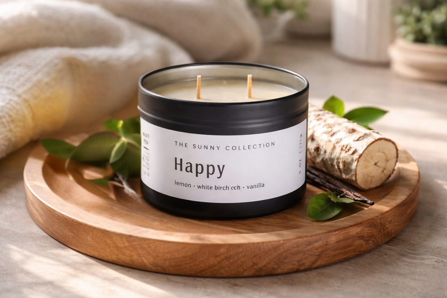 Scented candle labeled 'Happy' from The Sunny Collection on a wooden tray with leaves and birch logs.