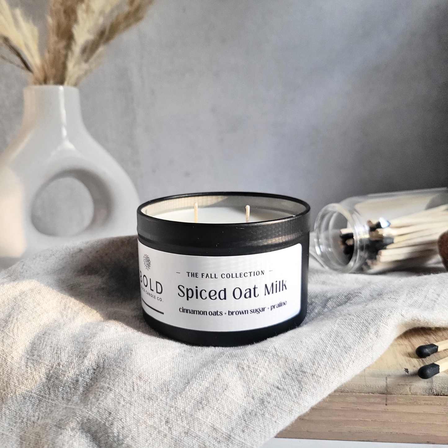 Candle labeled 'Spiced Oat Milk' from the 'The Fall Collection' on a textured surface with a neutral background.