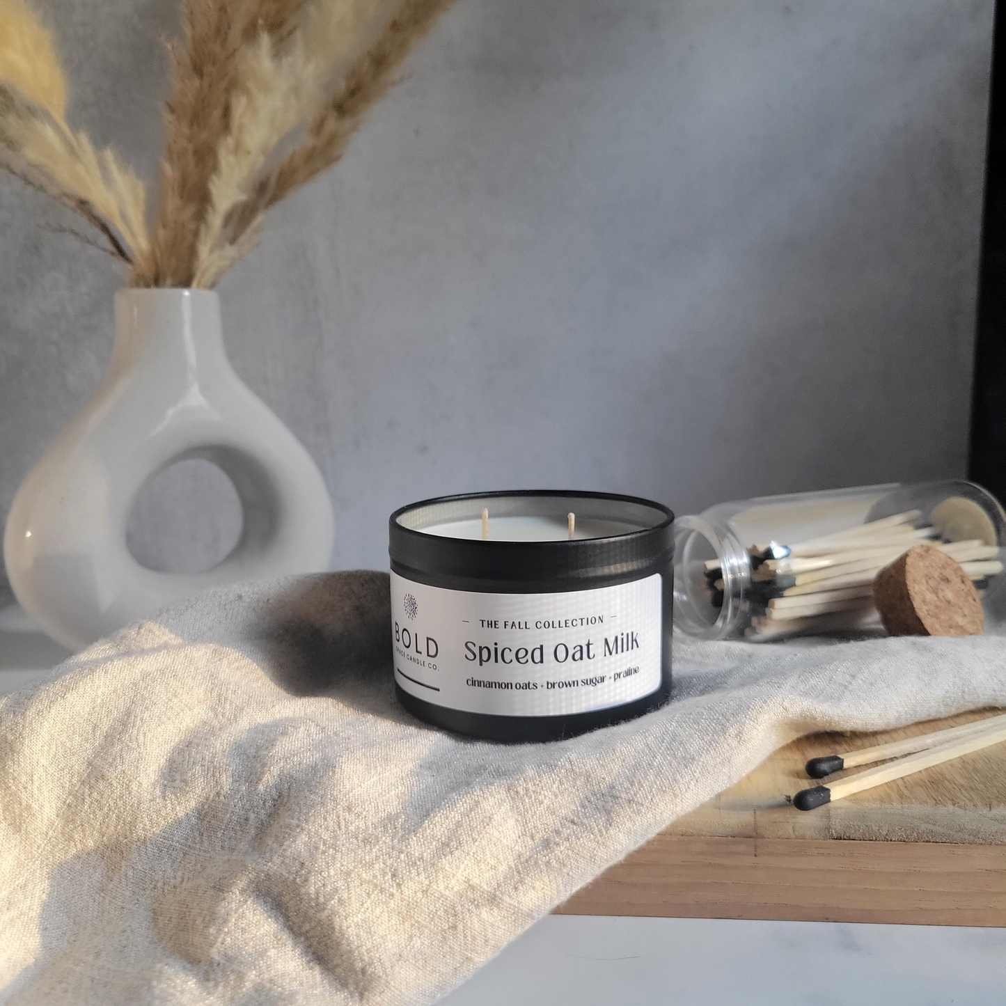 Candle labeled 'Spiced Oat Milk' on a textured surface with a vase and matches in the background.