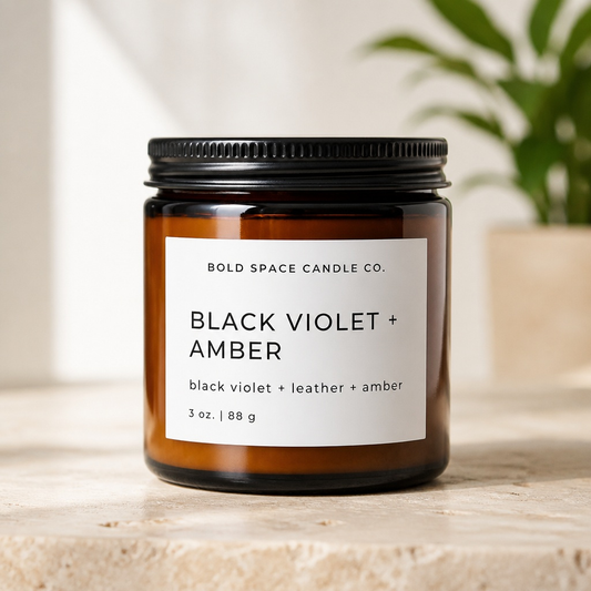 Amber candle jar with black lid and label on a light surface, plant in the background