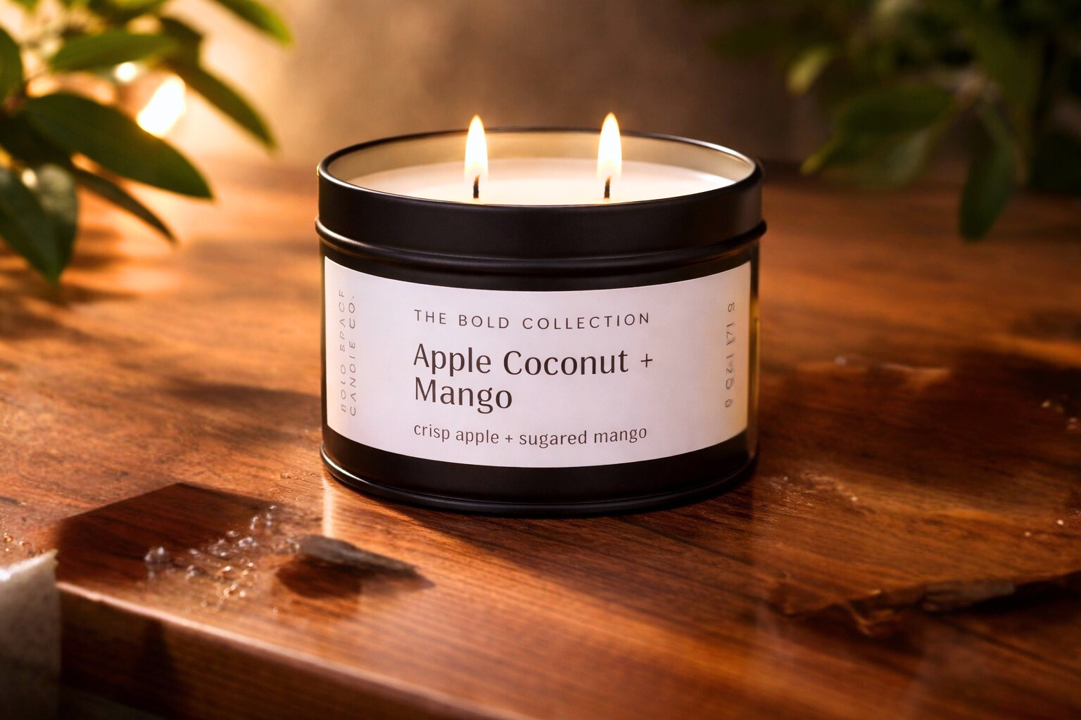 Candle labeled 'Apple Coconut + Mango' from 'The Bold Collection' on a wooden surface with blurred greenery in the background.