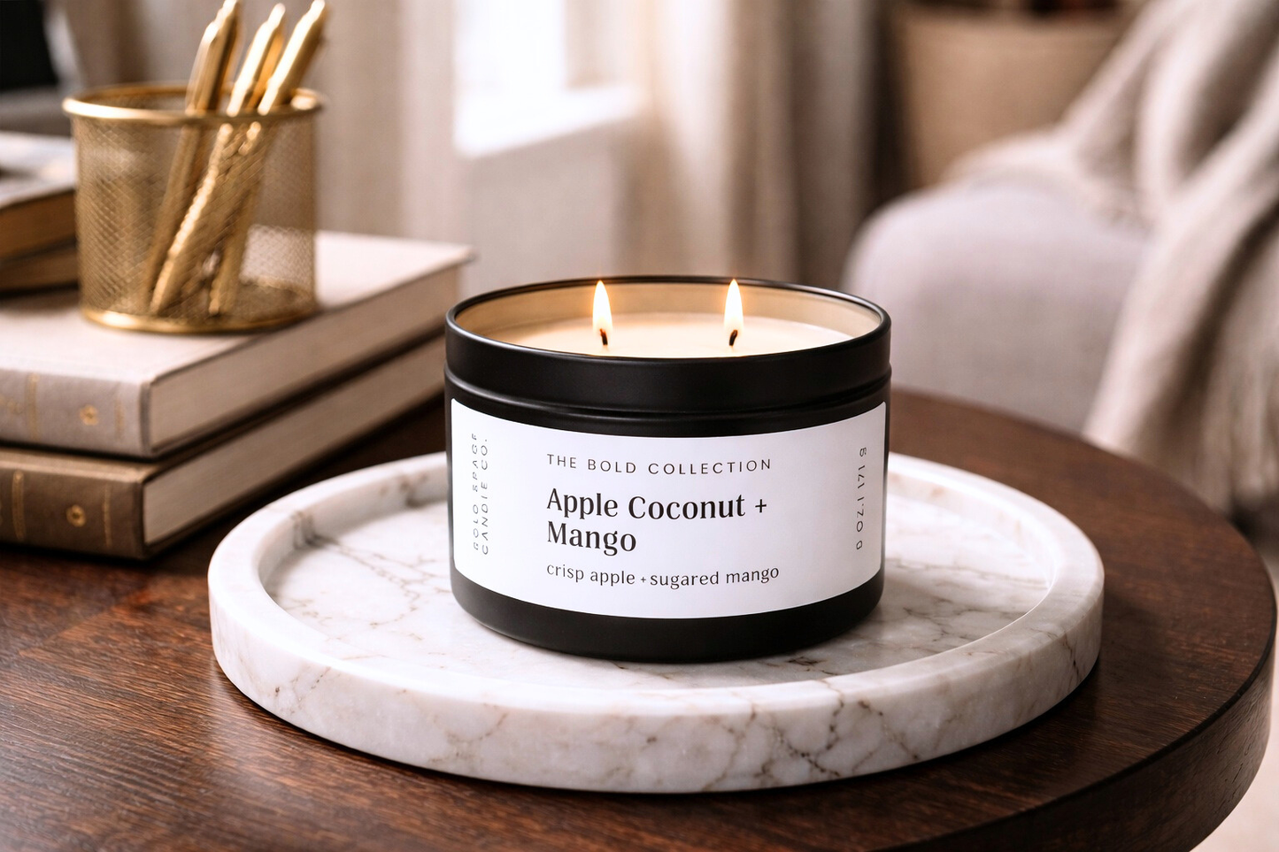 Candle labeled 'Apple Coconut + Mango' on a marble tray with books and a gold container in the background.