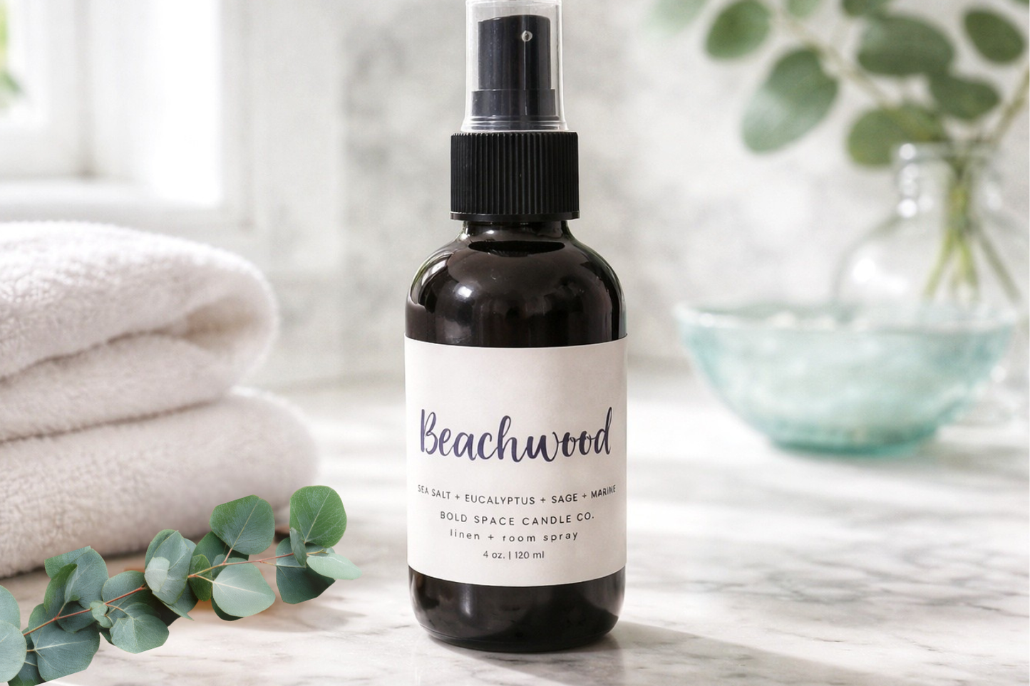 Bottle of Beachwood diffuser spray on a marble surface with eucalyptus leaves and towels in the background.