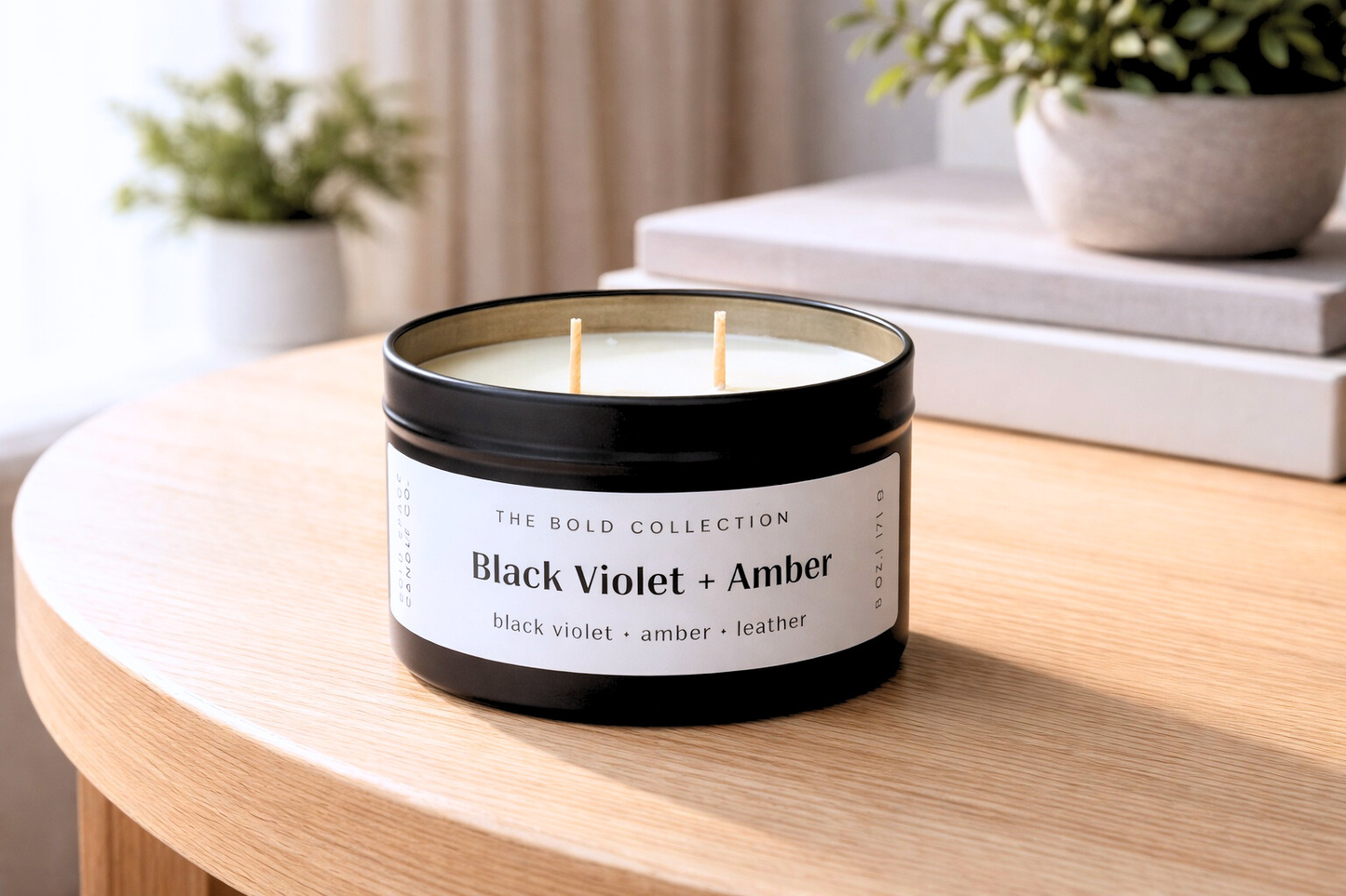 Candle in a black container labeled 'Black Violet + Amber' on a wooden surface with plants and books in the background.