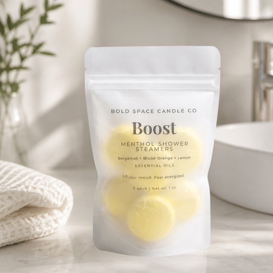 A white stand-up pouch of Bold Space Candle Co. Boost Menthol Shower Steamers sits on a light marble surface with a folded cream towel, a glass vase with greenery, and a softly blurred bathroom background.
