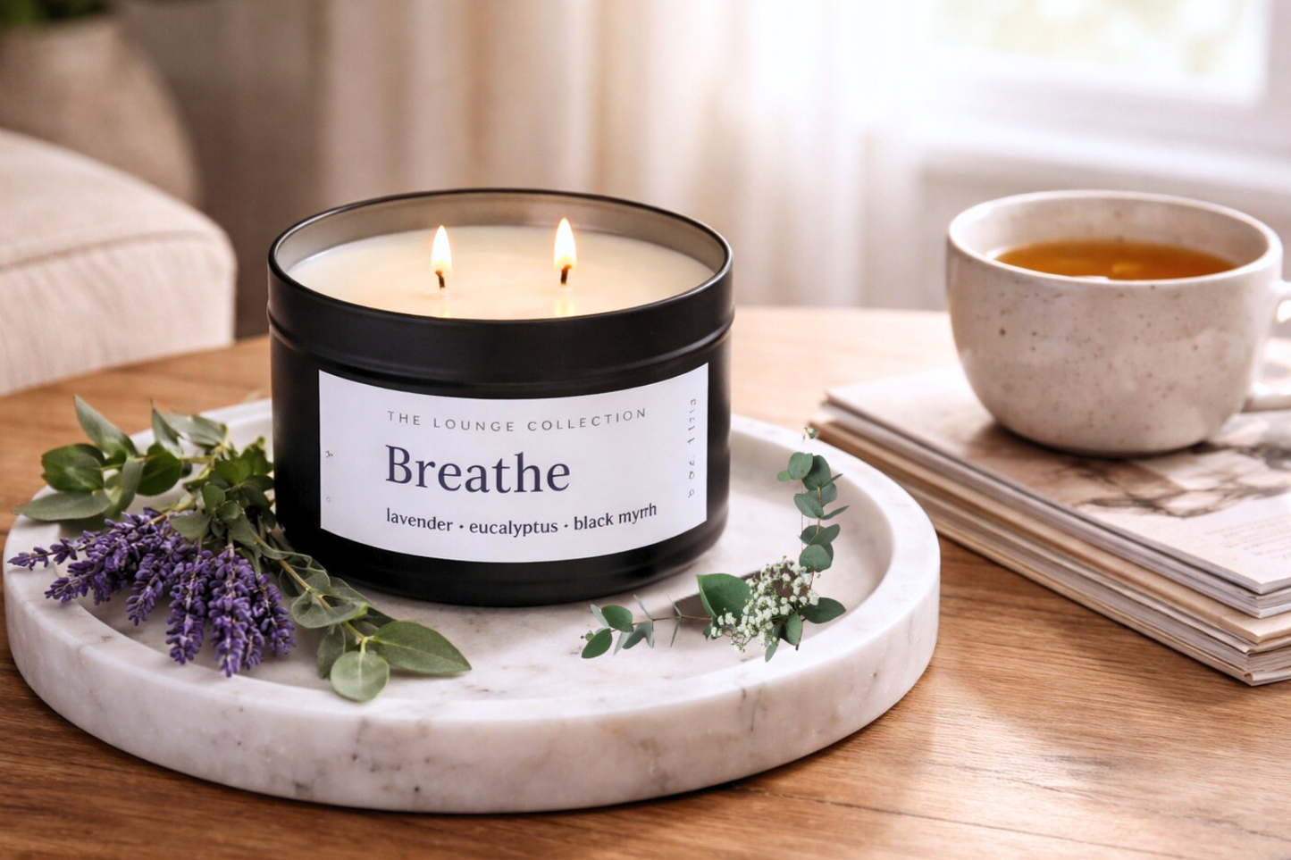 Candle labeled 'Breathe' on a marble tray with tea and flowers on a wooden table.
