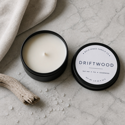 An open black candle tin showing creamy off-white wax, with the matching lid placed beside it on a light stone surface. The lid label reads 'DRIFTWOOD' and 'BOLD SPACE CANDLE CO.