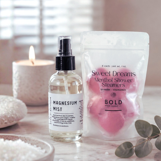 Bottle of magnesium mist and package of 'Sweet Dreams' shower steamers on a table with a candle and eucalyptus leaves.
