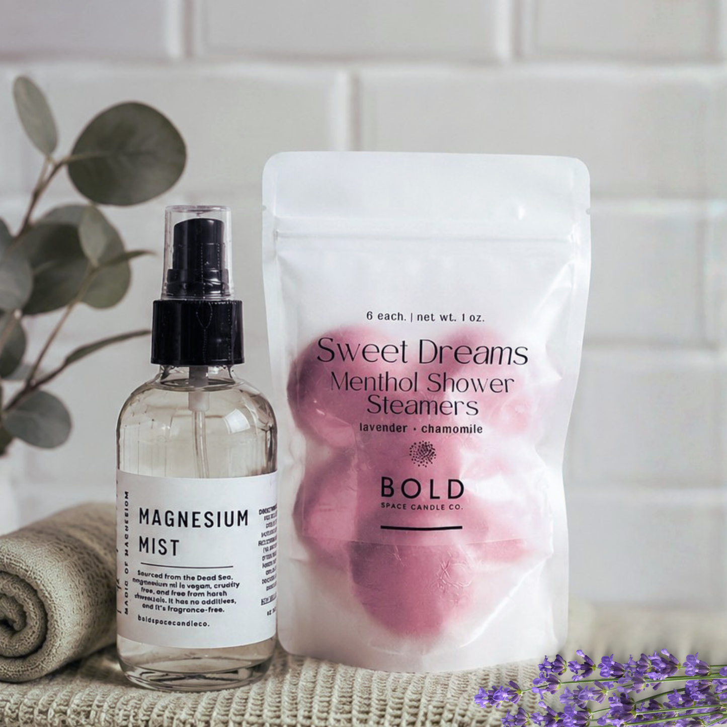 Bottle of magnesium mist and package of shower steamers on a surface with a plant and lavender in the background.