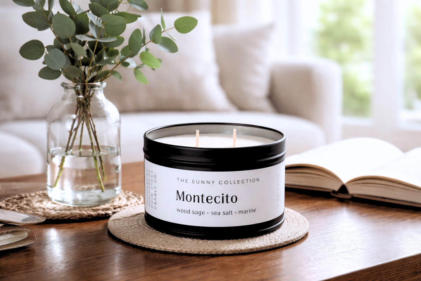 Candle labeled 'Montecito' from the Sunny Collection on a table with a vase of greenery and an open book in the background.