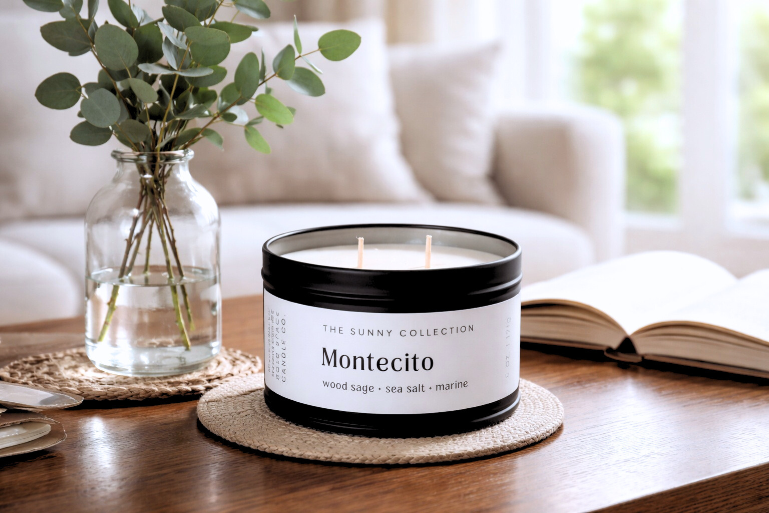 Candle labeled 'Montecito' from the Sunny Collection on a table with a vase of greenery and an open book in the background.
