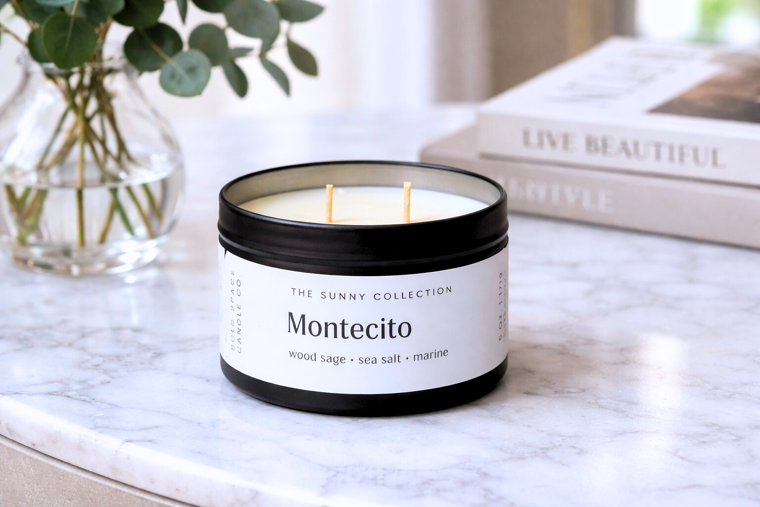 Candle in a container labeled 'Montecito' from 'The Sunny Collection' on a marble surface with books and a plant in the background.