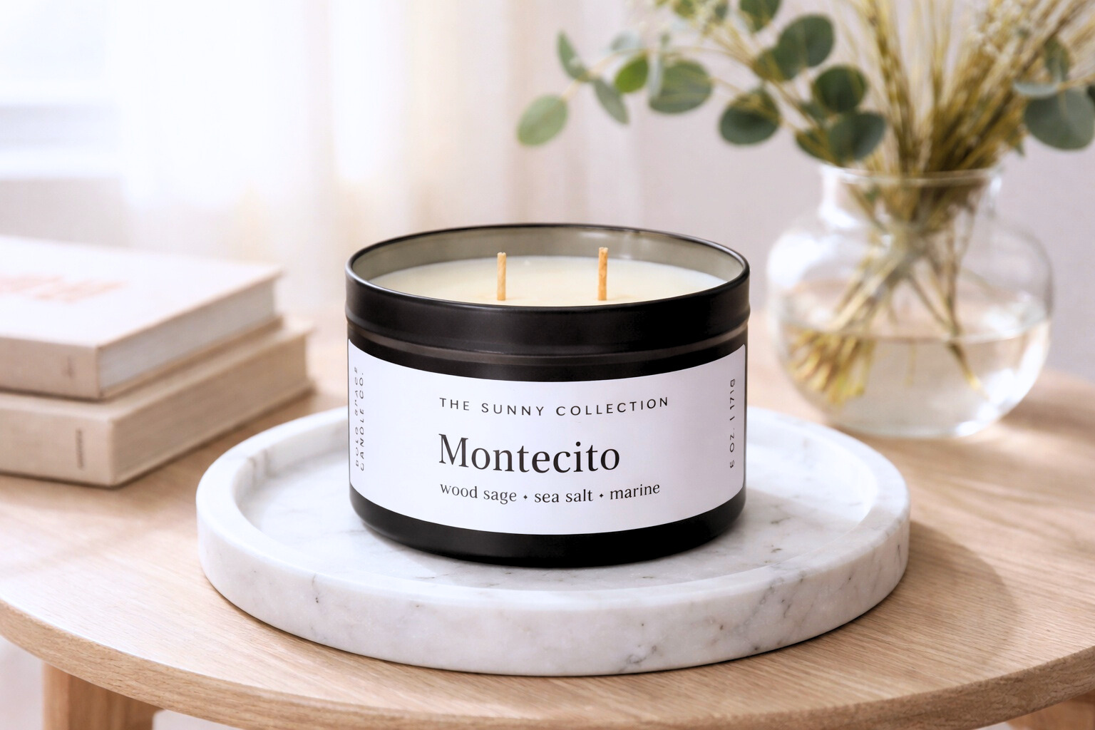Montecito candle from The Sunny Collection on a marble tray with books and a plant in the background.