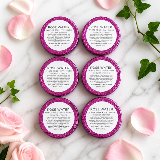 Six round rose colored foil shower steamers arranged in two columns on a white marble surface with pink rose petals and green leaves around them. The tins have bright pink glitter rims and white labels reading 'ROSE WATER' with product details below.