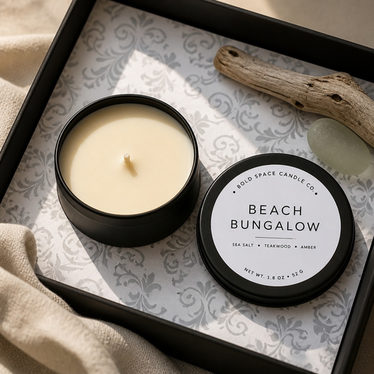 An open black tin candle and its matching lid labeled 'BEACH BUNGALOW' by Solid Space Candle Co., arranged in a black gift box with patterned liner, beige fabric, driftwood, and a green stone.