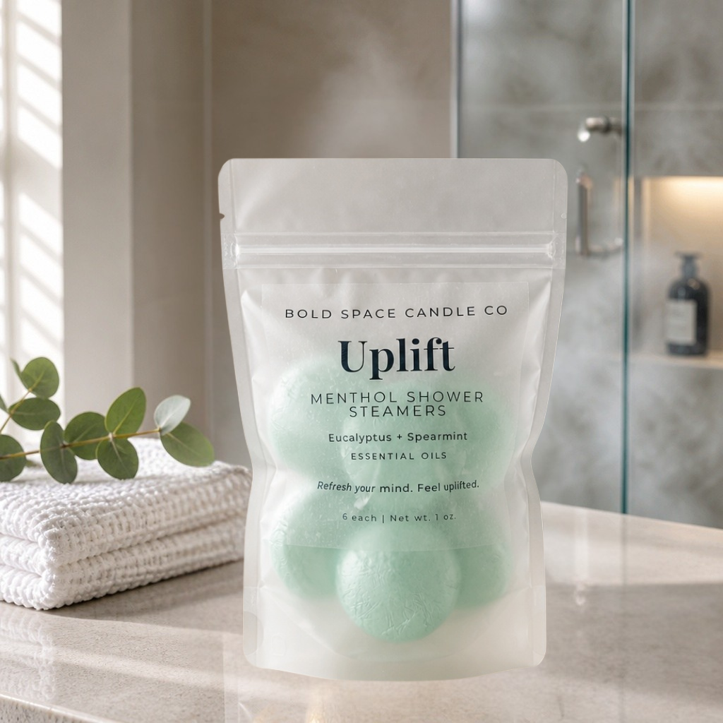 A stand-up pouch of Bold Space Candle Co. Uplift Menthol Shower Steamers sits on a bathroom counter with a folded towel and eucalyptus leaves nearby.