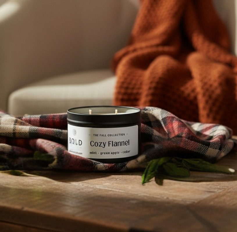 Candle labeled 'Cozy Flannel' on a wooden surface with plaid fabric and a blanket in the background